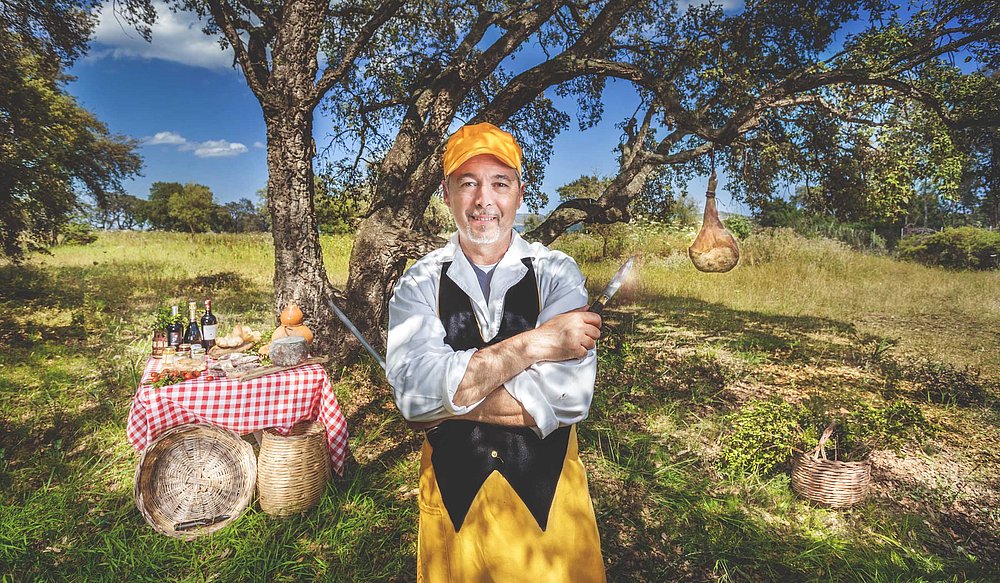 Uomo sorridente in abiti da cuoco, con cappello giallo, sta in un prato sotto un albero. Accanto a lui, un tavolo apparecchiato con cibo e bevande. Il paesaggio è verde e soleggiato, creando un'atmosfera accogliente e rustica.