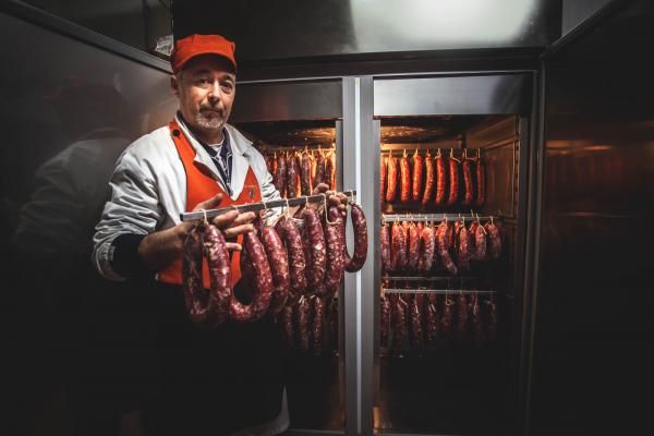 A person wearing a red cap and apron stands in front of a refrigerator, holding a rack of sausages. The fridge contains hanging sausages arranged neatly, showcasing a variety of cured meats. The setting appears to be a meat shop or deli.